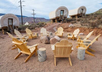 Outdoor wooden chairs facing covered wagons, desert setting.